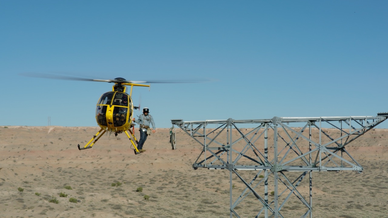 Lineman riding on the skid of a yellow helicopter hovering beside a high-voltage tower, from the Boot Barn Higher Ground commercial