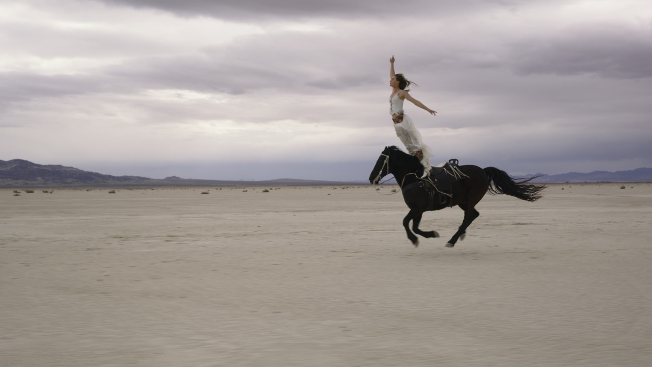 Trick rider in a white dress standing with arms outstretched on a galloping black horse across a desert salt flat, from the Boot Barn commercial